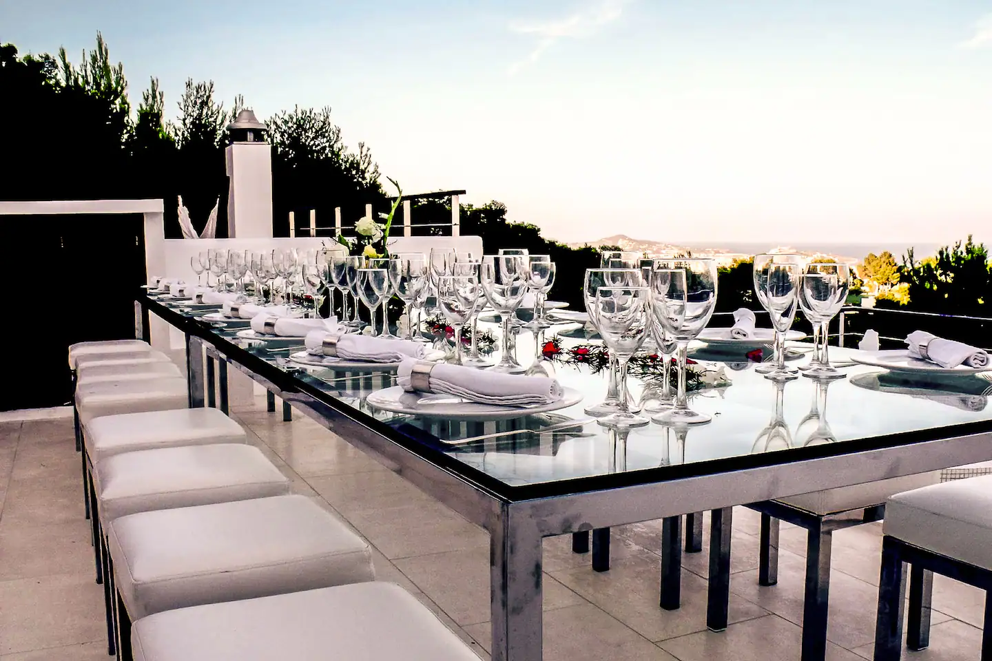 An inviting outdoor dining room set amidst a beautifully landscaped garden, with a long glass table and comfortable chairs.